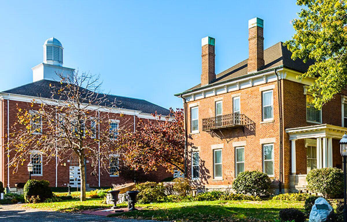 Brick campus buildings with a small balcony, trees, and a lawn under a clear blue sky.