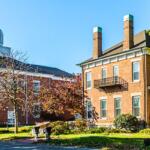 Brick campus buildings with a small balcony, trees, and a lawn under a clear blue sky.