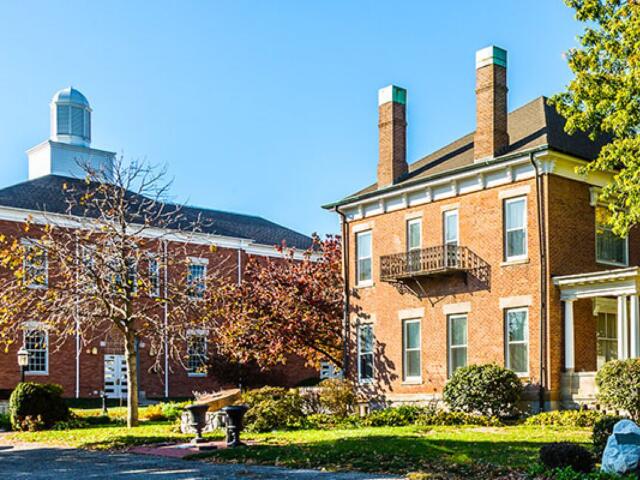 Brick campus buildings with a small balcony, trees, and a lawn under a clear blue sky.