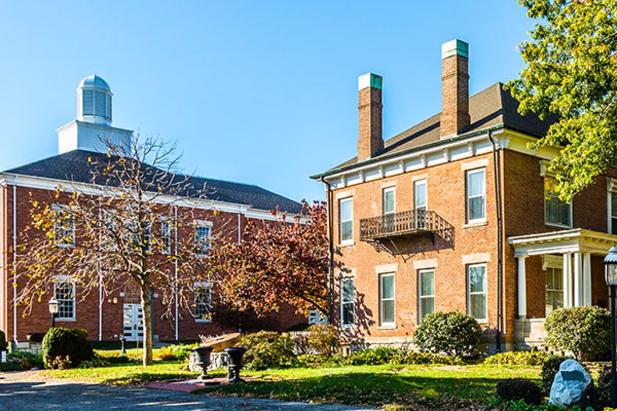 Brick campus buildings with a small balcony, trees, and a lawn under a clear blue sky.