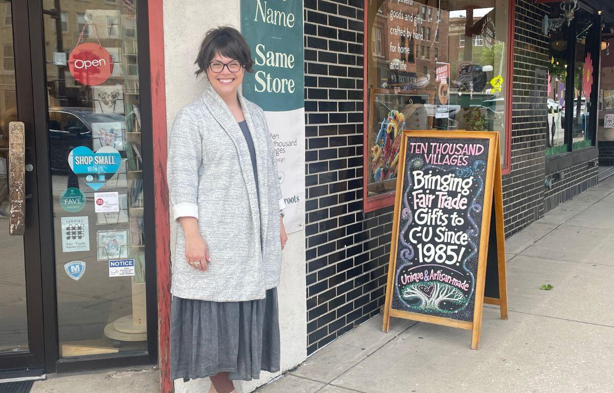 Smiling woman in a gray coat and glasses stands outside a storefront with a chalkboard sign promoting fair trade gifts since 1985.