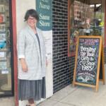 Smiling woman in a gray coat and glasses stands outside a storefront with a chalkboard sign promoting fair trade gifts since 1985.