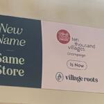Sign announcing a rebrand: 'New Name Same Store' on a dark panel; right panel features the 'ten thousand villages' logo and the message 'Is Now Village Roots' for Champaign.