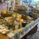 Table in a gift shop full of handmade crafts—ceramics, candles, small plants, cards and books on a floral tablecloth.