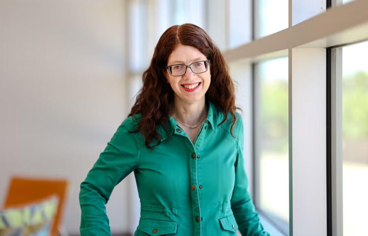 Smiling woman with glasses and curly red hair standing by a large window in a bright, modern interior, wearing a teal button-down shirt.