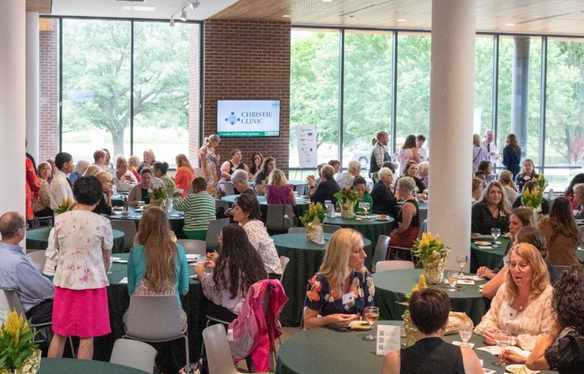 People gather at a daytime conference or reception around round tables with green tablecloths and yellow flower centerpieces; a Christie Clinic screen is visible in the background.