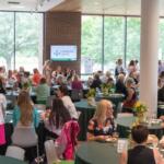 People gather at a daytime conference or reception around round tables with green tablecloths and yellow flower centerpieces; a Christie Clinic screen is visible in the background.