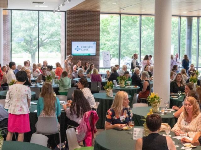 People gather at a daytime conference or reception around round tables with green tablecloths and yellow flower centerpieces; a Christie Clinic screen is visible in the background.