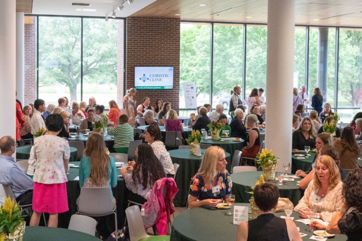 People gather at a daytime conference or reception around round tables with green tablecloths and yellow flower centerpieces; a Christie Clinic screen is visible in the background.