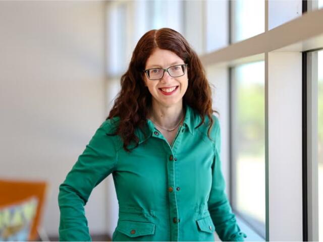 Smiling woman with glasses and curly red hair standing by a large window in a bright, modern interior, wearing a teal button-down shirt.
