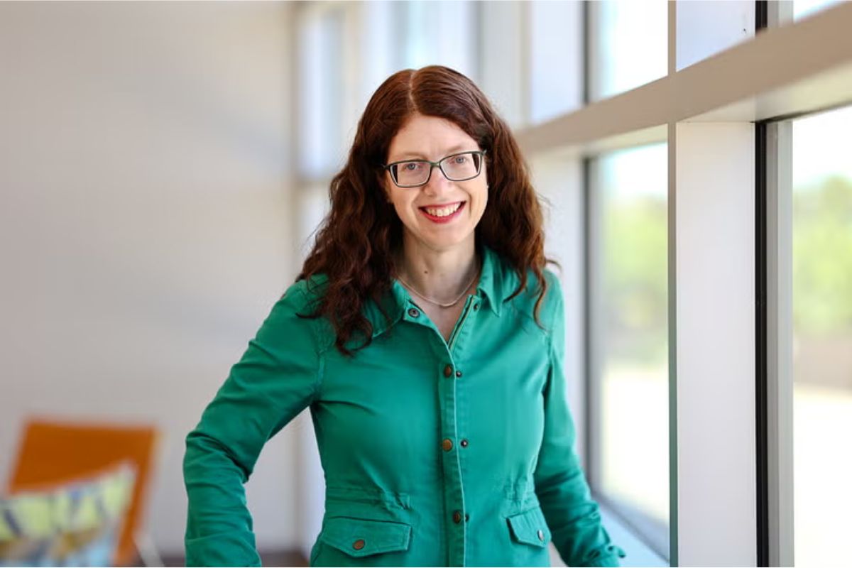 Smiling woman with glasses and curly red hair standing by a large window in a bright, modern interior, wearing a teal button-down shirt.