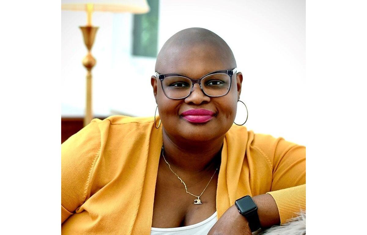 Smiling bald woman with glasses and hoop earrings in a mustard cardigan, wearing a necklace and a black smartwatch, seated indoors.