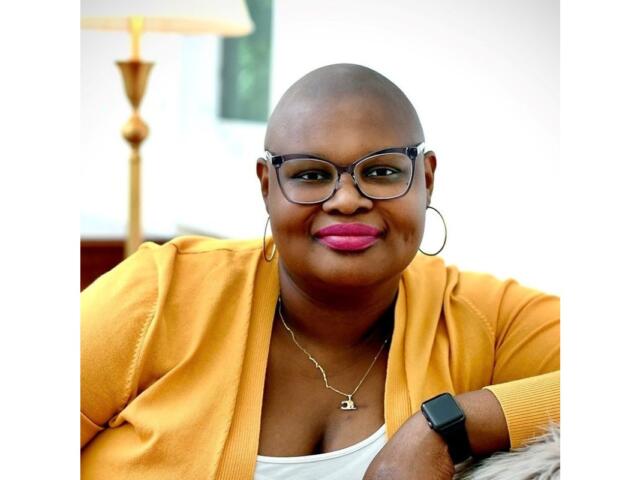 Smiling bald woman with glasses and hoop earrings in a mustard cardigan, wearing a necklace and a black smartwatch, seated indoors.
