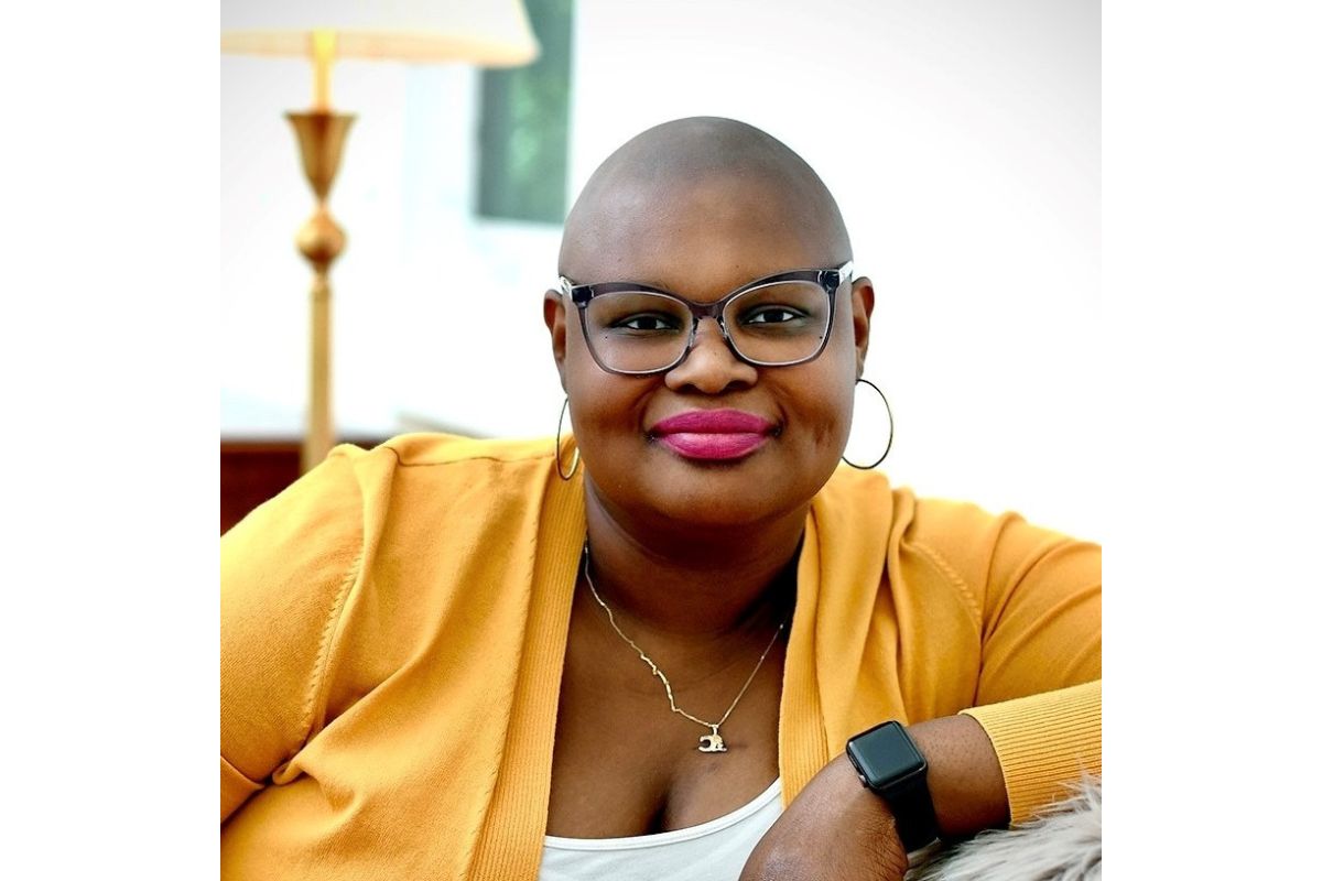 Smiling bald woman with glasses and hoop earrings in a mustard cardigan, wearing a necklace and a black smartwatch, seated indoors.