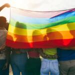 Group of people holding a large rainbow pride flag, embracing in a sunny outdoor setting, symbolizing LGBTQ+ pride and solidarity.