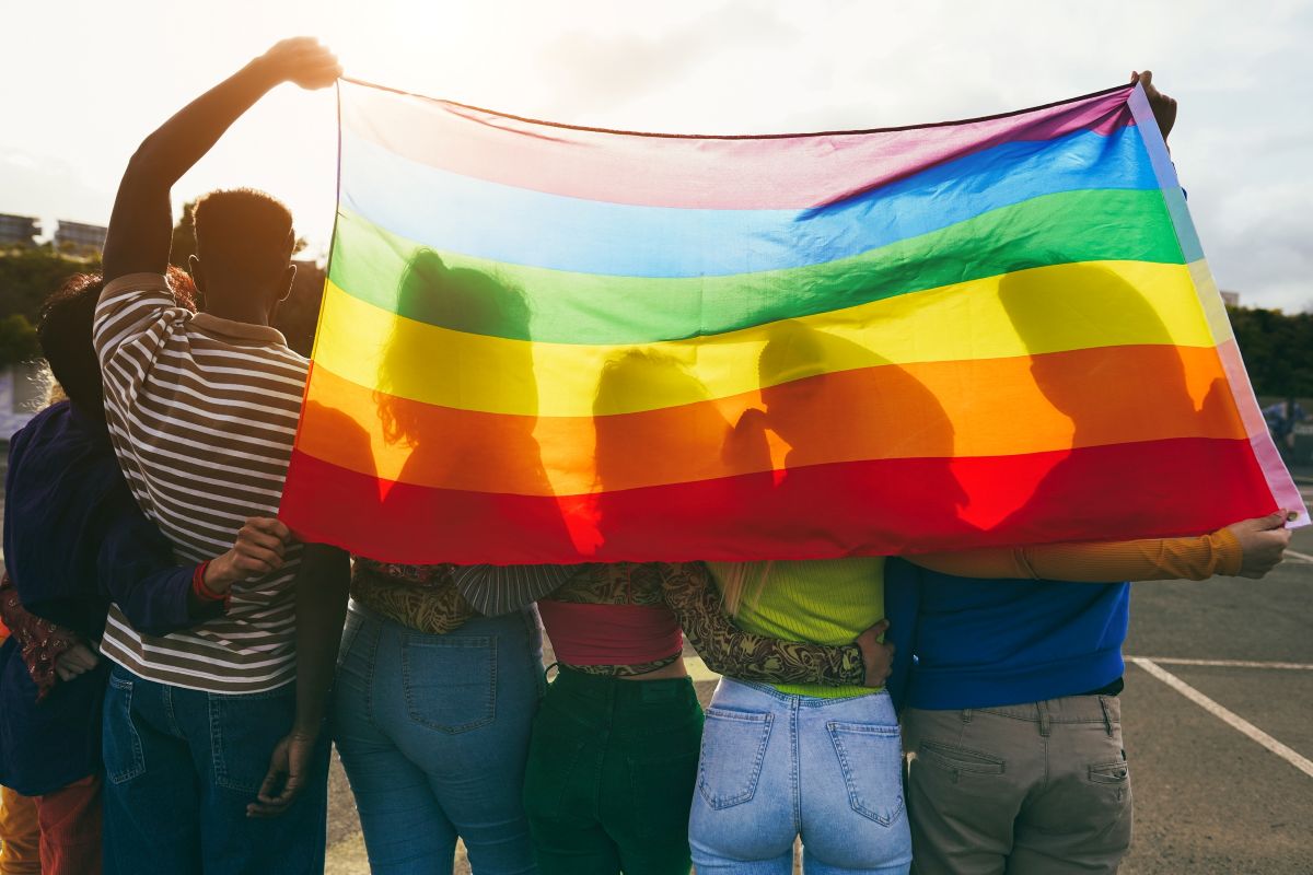 Group of people holding a large rainbow pride flag, embracing in a sunny outdoor setting, symbolizing LGBTQ+ pride and solidarity.