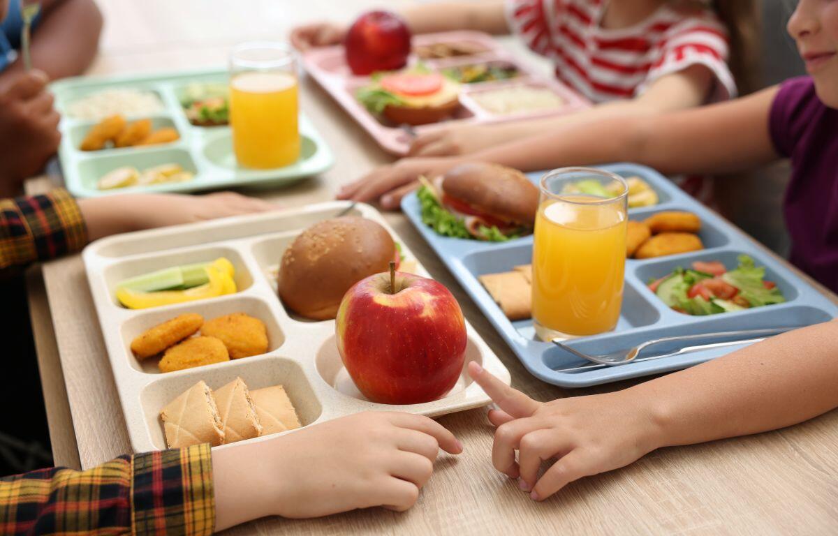 Children share a cafeteria lunch at a table with multi-compartment trays containing an apple, sandwich, nuggets, cucumber, and juice cups in front of them.