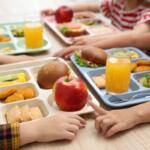 Children share a cafeteria lunch at a table with multi-compartment trays containing an apple, sandwich, nuggets, cucumber, and juice cups in front of them.