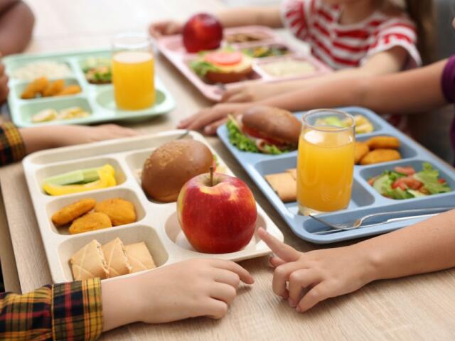 Children share a cafeteria lunch at a table with multi-compartment trays containing an apple, sandwich, nuggets, cucumber, and juice cups in front of them.