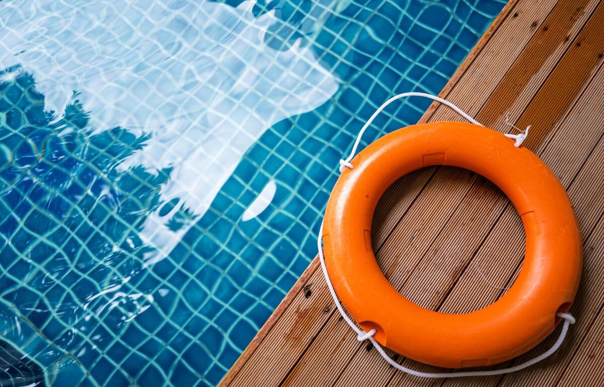 Orange lifebuoy with white rope on a wooden pool deck beside a blue tiled pool surface.