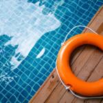 Orange lifebuoy with white rope on a wooden pool deck beside a blue tiled pool surface.