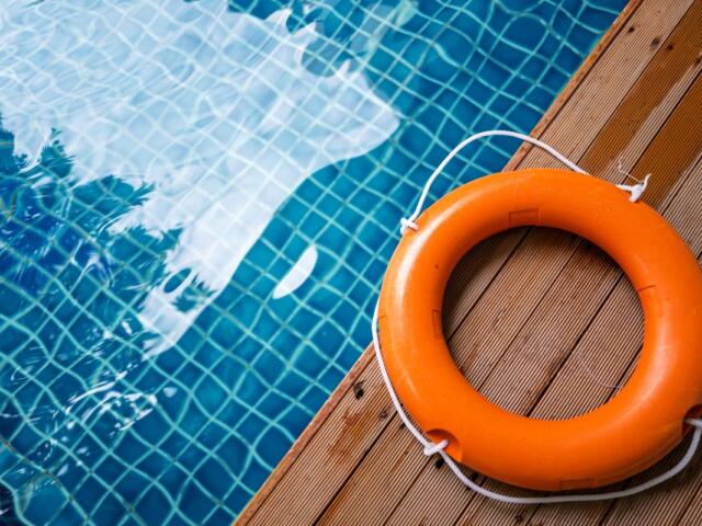 Orange lifebuoy with white rope on a wooden pool deck beside a blue tiled pool surface.