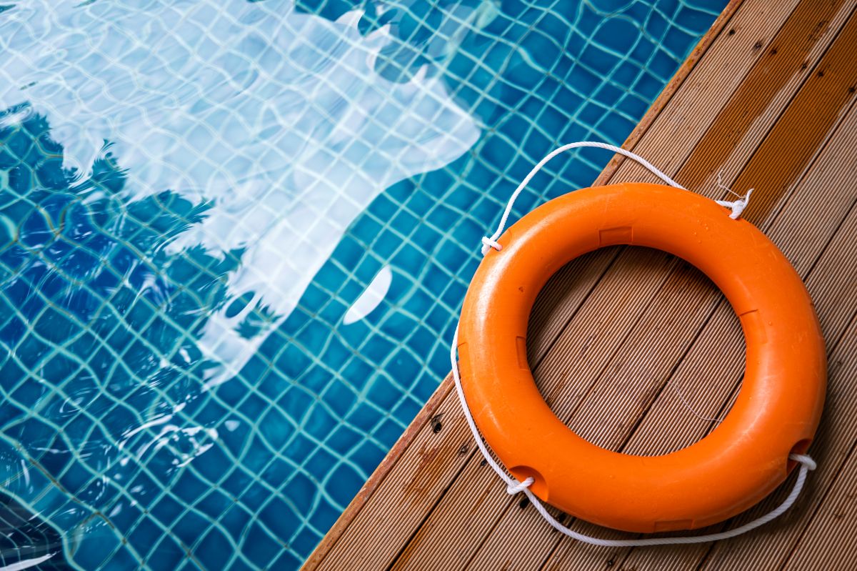 Orange lifebuoy with white rope on a wooden pool deck beside a blue tiled pool surface.