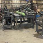 Industrial workshop with metalworking tables, tools, and a green gas cylinder on a wheeled cart; cluttered backdrop of equipment and wiring.