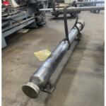Long metal cylindrical pipe on wheeled supports in an industrial workshop, secured with a lifting strap close to the pipe end.