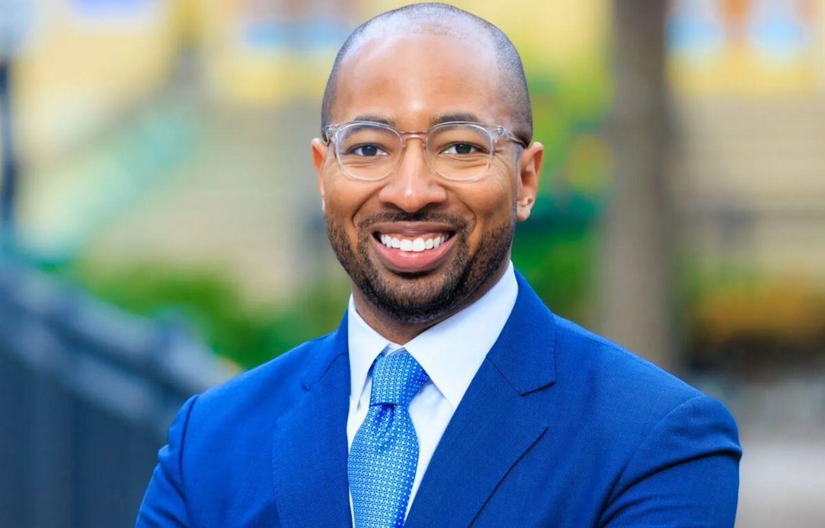 Professional headshot of a smiling man in a blue suit and tie, outdoor background blurred behind him.