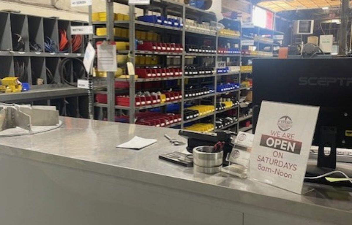Checkout counter at a hardware store; organized shelves with red, yellow, and blue bins behind; sign reads 'We are open on Saturdays 8am–Noon'