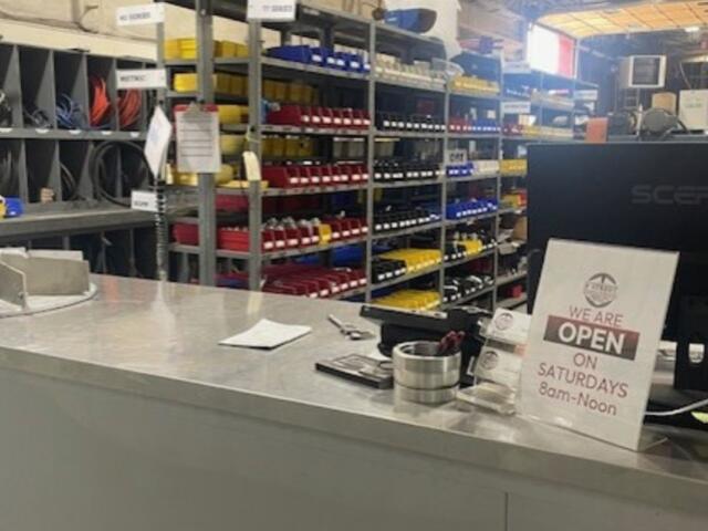 Checkout counter at a hardware store; organized shelves with red, yellow, and blue bins behind; sign reads 'We are open on Saturdays 8am–Noon'