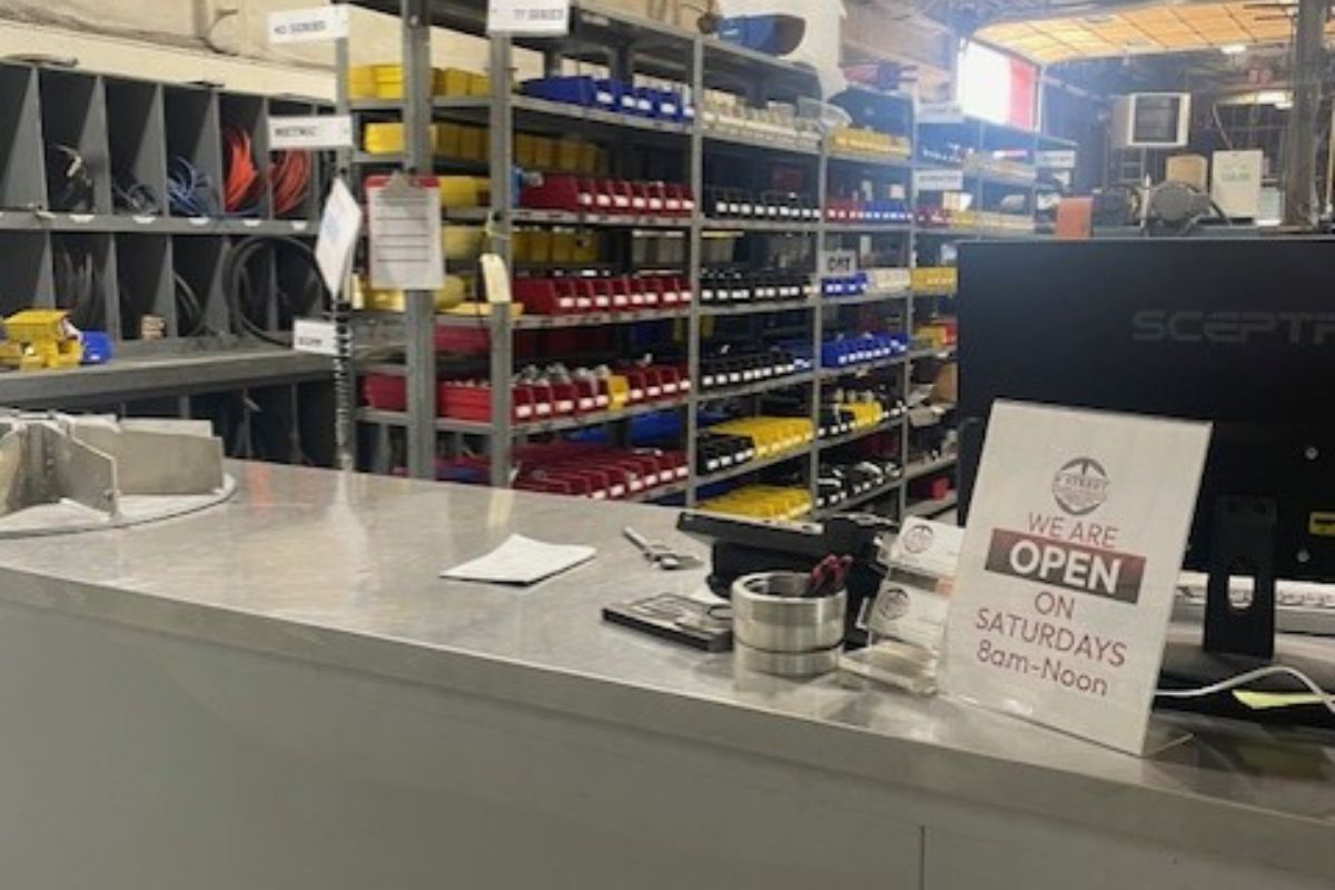 Checkout counter at a hardware store; organized shelves with red, yellow, and blue bins behind; sign reads 'We are open on Saturdays 8am–Noon'