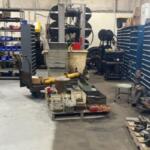 Cluttered industrial workshop with shelves, bins, and scattered parts on pallets; tires and equipment in the background.