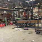 Industrial workshop scene with heavy machinery, metal frames, tools, and an overhead crane in a cluttered shop.