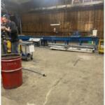 Worker in safety glasses welding a metal piece beside a blue fabrication table in an industrial workshop.