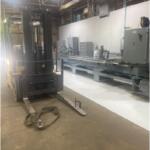 Forklift parked in an industrial workspace next to a long metal conveyor with control panels and machinery along a white wall. The forks are lowered to the floor and a strap lies nearby on the dusty concrete surface.