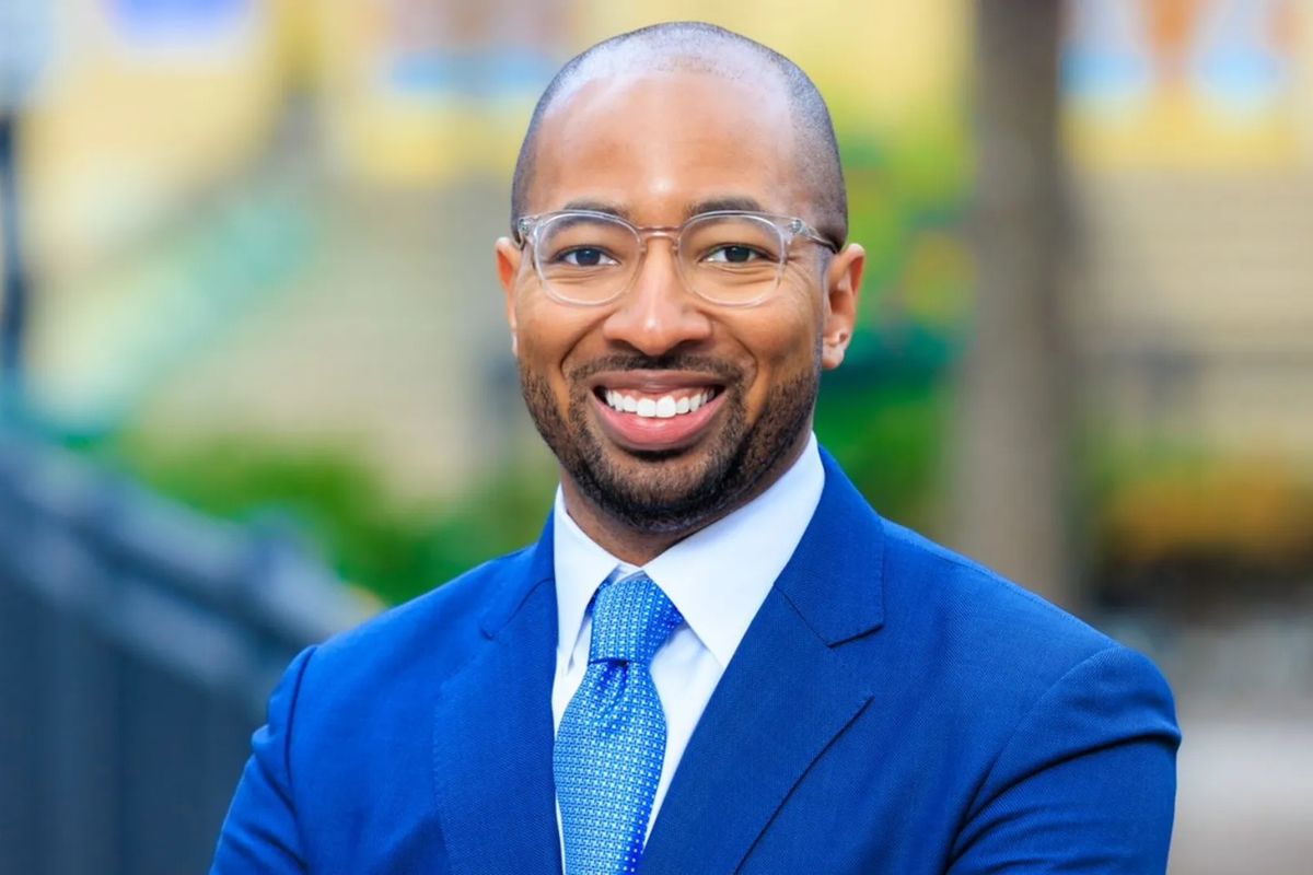 Professional headshot of a smiling man in a blue suit and tie, outdoor background blurred behind him.